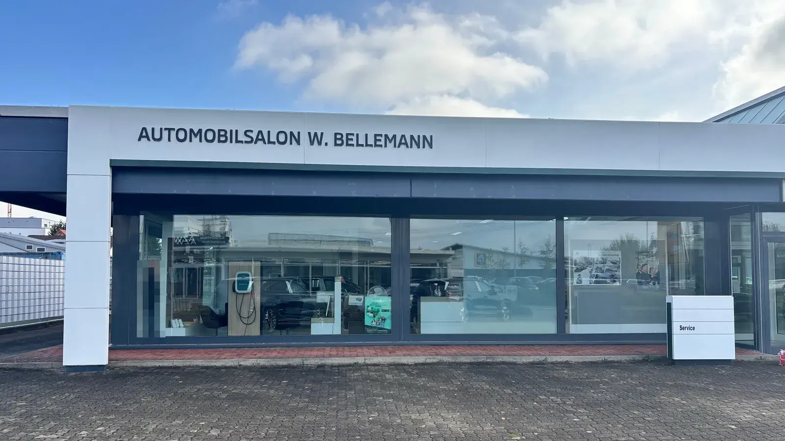 Automobilsalon Bellemann in Wiesloch: Gebäudefront mit dem Schriftzug Automobilsalon W. Bellemann, Schaufenster mit Fahrzeugen und Ladesäule, rechts die Stele mit der Aufschrift Service